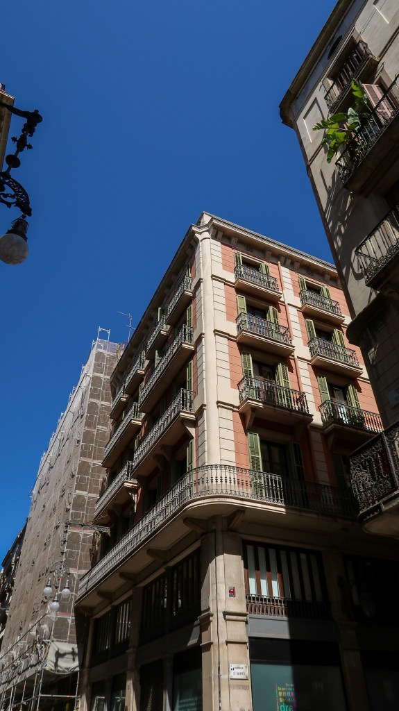 An image of a corner of a building in a block. It is a six story building in orange with white brick trim. Each rectangle window is green and has a balcony. There is a another building in the right corner of the image. The sky above is blue. 
