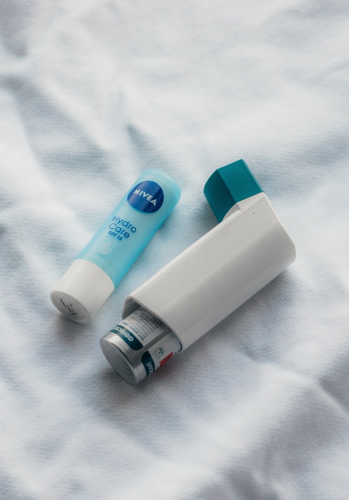 A white inhaler with a blue lid laid on a creased white sheet. To the left of the inhaler is a blue chapstick.