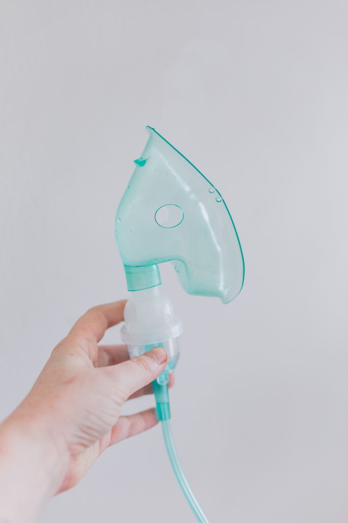 A teal oxygen mask being held up by a white hand against a light grey background. 