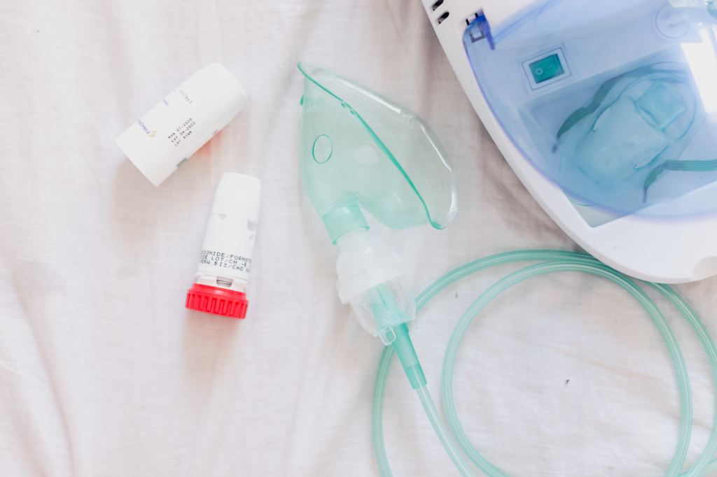 Two white inhalers, one with a red base, on a white sheet. To the right of the inhalers is a teal oxygen mask with a teal tube. To the right of that is a white box with a clear blue lid. 