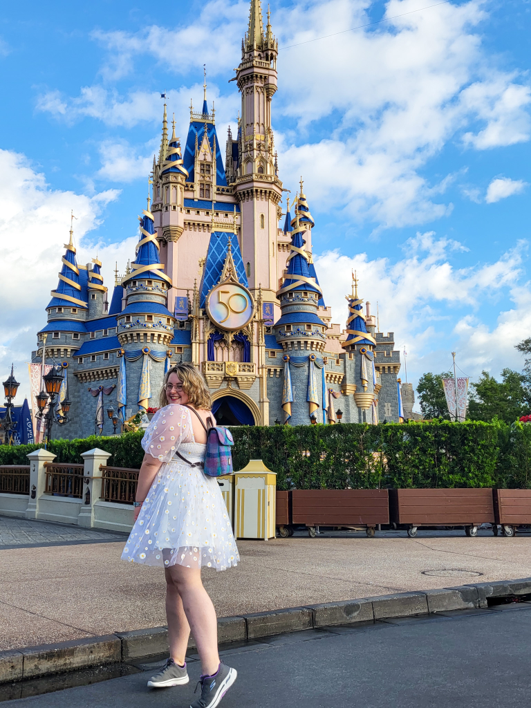 A picture of Cinderella's Castle, in all its blue and pastel pink glory, against a blue sky with white clouds. Rosie, a white woman with short wavy blonde hair, stands in front of the castle, looking over her shoulder. She is wearing a white dress, a blue backpack, and grey trainers. 