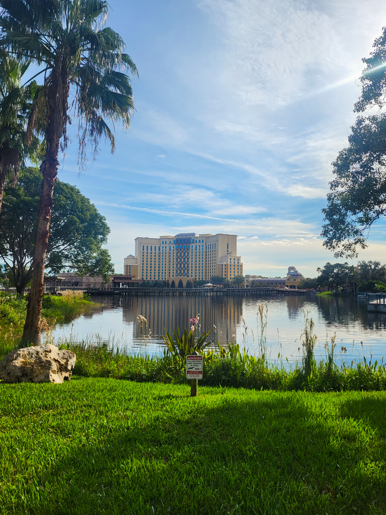A yellow/orange tower hotel in the distance in front of  blue lake and against a blue and white sky. In front of the lake is green grass, water plants, and a palm tree. 