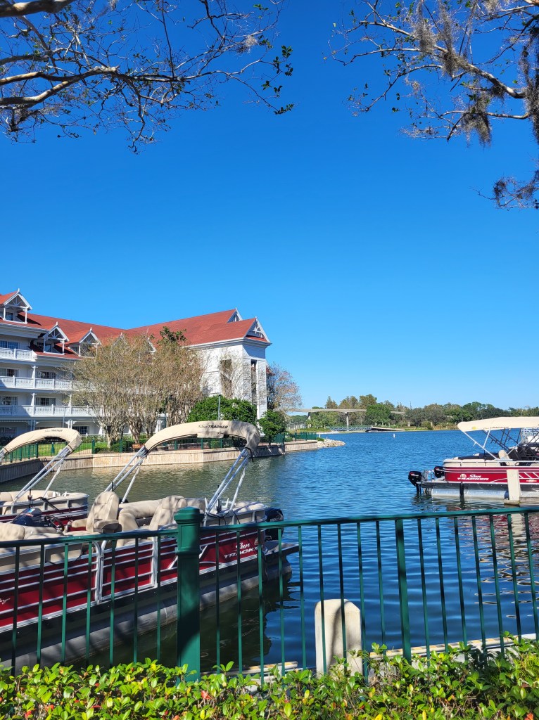 The boat rental at Grand Floridian Resort. There is a lake with red boats, in front of a white building with a red roof, and greenery. 