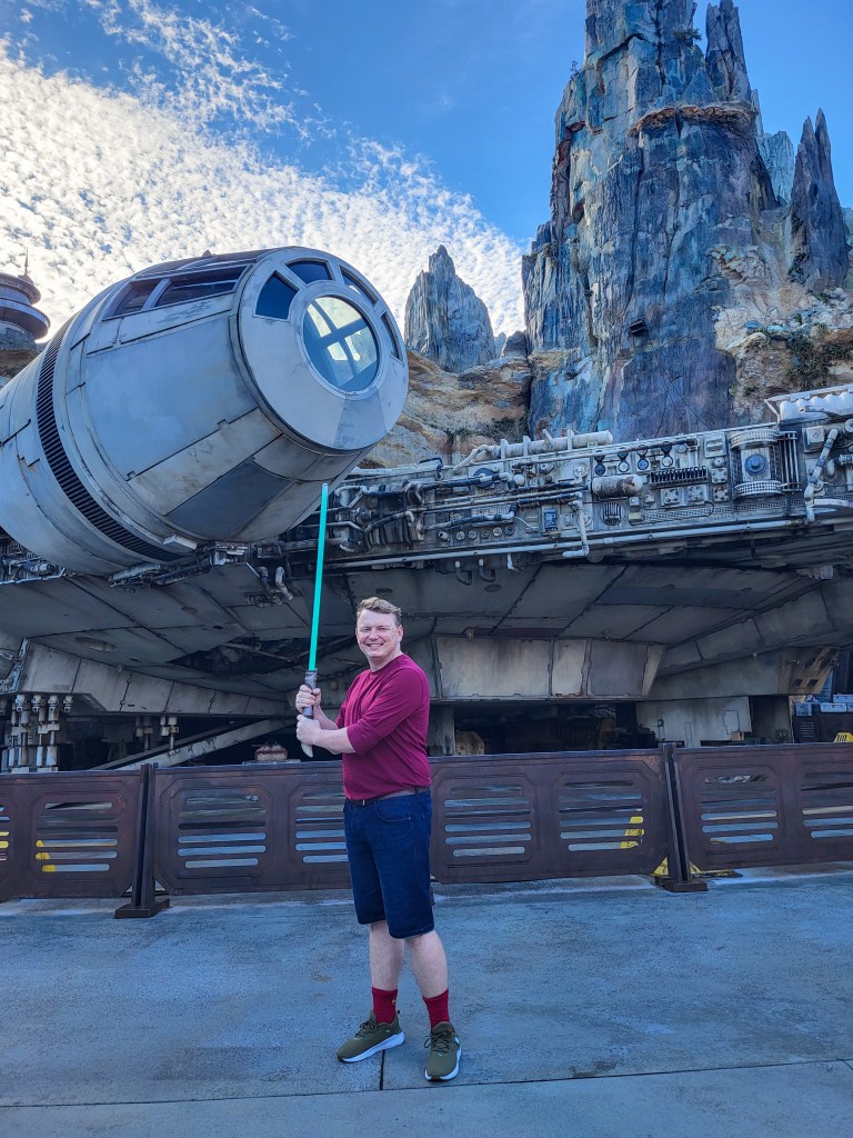 Rory (a white man with short brown hair, a red long sleeved tee, blue shorts, and trainers) is standing in front of the Millennium Falcon. He is holding a green lightsaber.