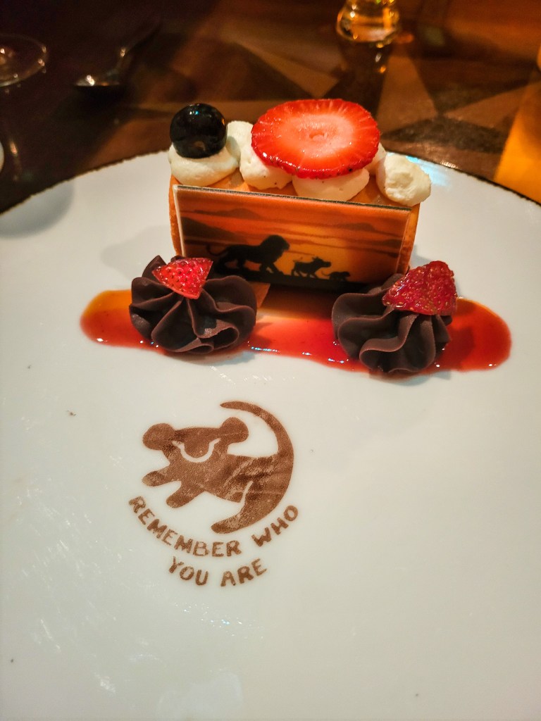A dessert on a white plate. It is a white brownie with a sunset from The Loin King on it, topped with fruit, and chocolate cream. There is a chocolate dusting that reads 'remember who you are' on it