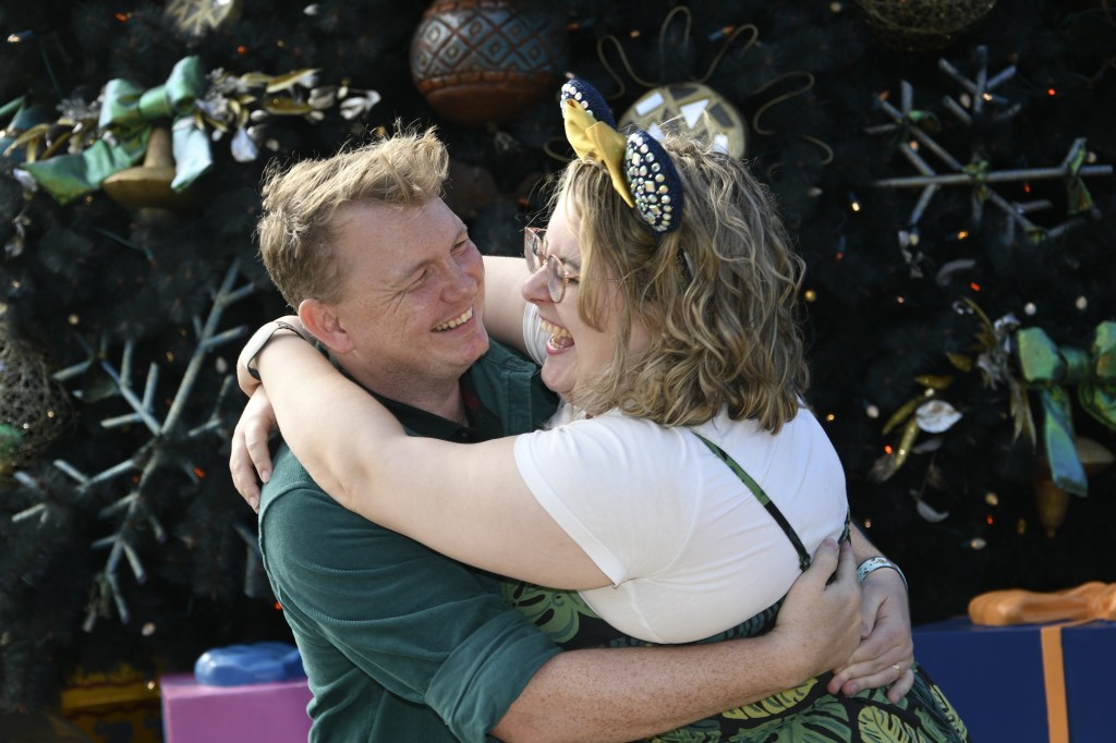 A couple embracing and laughing. A white man is on the left with light brown, short, wavy hair, and he is smiling. He is wearing a dark green shirt. A white women is on the right, with blonde wavy shoulder length hair , glasses, and is wearing blue Mickey ears with a yellow bow. She is  wearing a white tee and a green leaf print dress. They are infront of a green tree background.