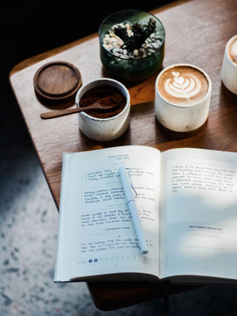 A notebook with black writing in it on a brown table. There is a plant, coffee in a white mug, and a sugar pot above it.