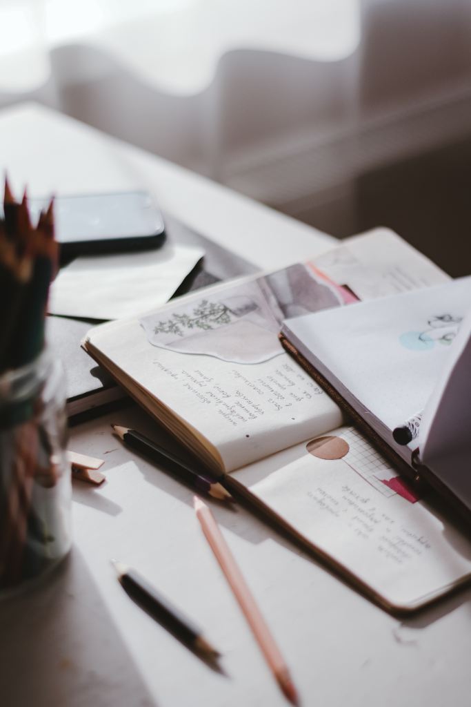Two artist's journals, open, on top of each other. There are pencils on the desk and a jar of pencils.