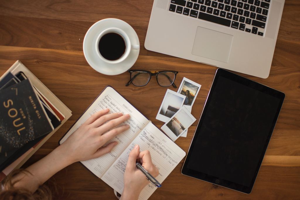 A white hand on top of a notebook, another hand writing in the notebook. A stack of books to the left; coffee, glasses, MacBook and polaroid pictures above; a black notebook to the right. This all rests on a wooden table.