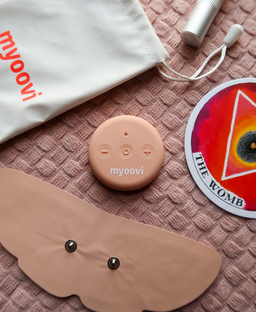 Items on a pink blanket. In the middle is a pale pink round object with buttons and says 'myoovi'. Above it is a white bag with 'myoovi' in orange. There is the lid of a concealer above and then a card on the right. The card is circular, red, and says 'the womb'. Below the round object is a pale pink butterfly pad.  