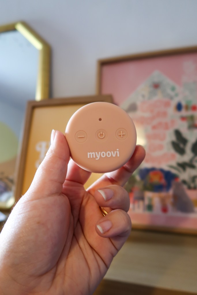 A pale pink round object with buttons and that says 'myoovi', being held by a white person's hand. Behind it, and out of focus, are framed art prints. 