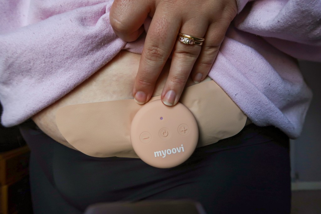 The circular myoovi object attached to the pad and on someone's stomach. A white hand touches the pale pink object; the hand is wearing two gold rings and also holding up a pink jumper. 
