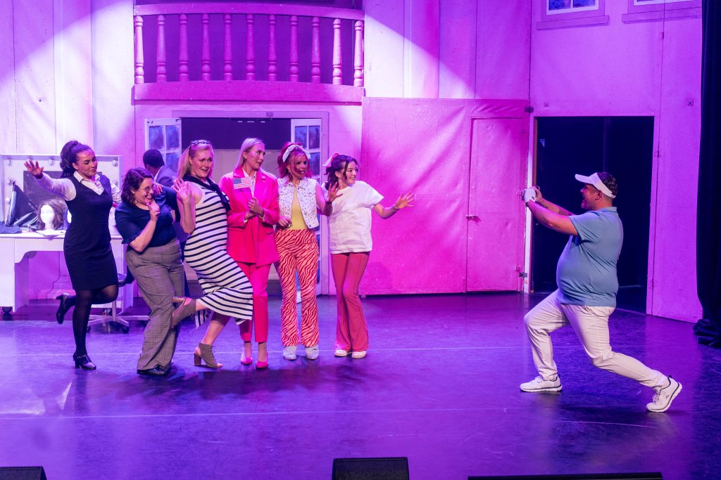 A picture of six women posing on stage whilst a man takes a photo, also on stage. They are against a pink background. 
