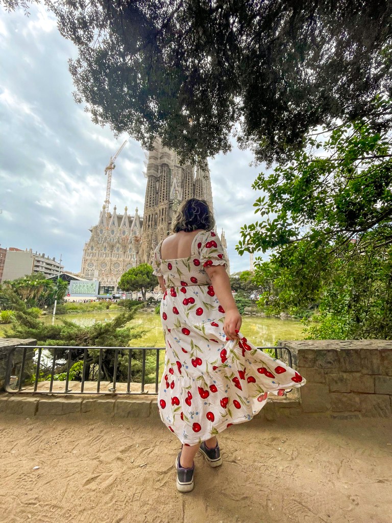 An image of Rosie stood facing La Sagrada Familia. She is a white woman with a wavy brown bob, and is twirling her dress. The dress is cream, with a cherry print with blue flowers. There is a green lake in front of her, and the sky is blue grey behind the cathedral. 