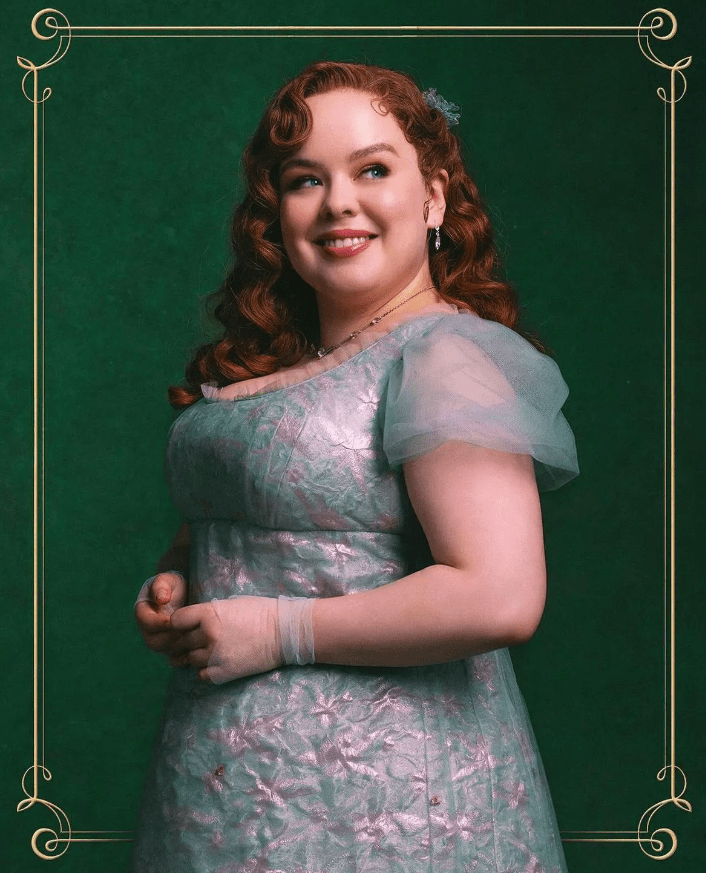 A portrait style photo of Nicola Coughlan as Penelope Featherington. She is a petite and rounded white woman, with wavy red long hair, and she is looking off to the right of the image, smiling. She has gentle make up on and is wearing a pale blue gown with tulle short sleeves. She is gently clasping her hands in front of her. The background of the image is dark green and surrounded by a gold trim. 