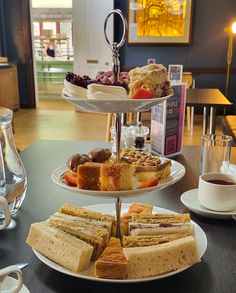 A triple stack afternoon tea with sandwiches' on the bottom, cakes in the middle, and sweet treats/scones on the top. This is on a black top table and you can see a cafe behind. 