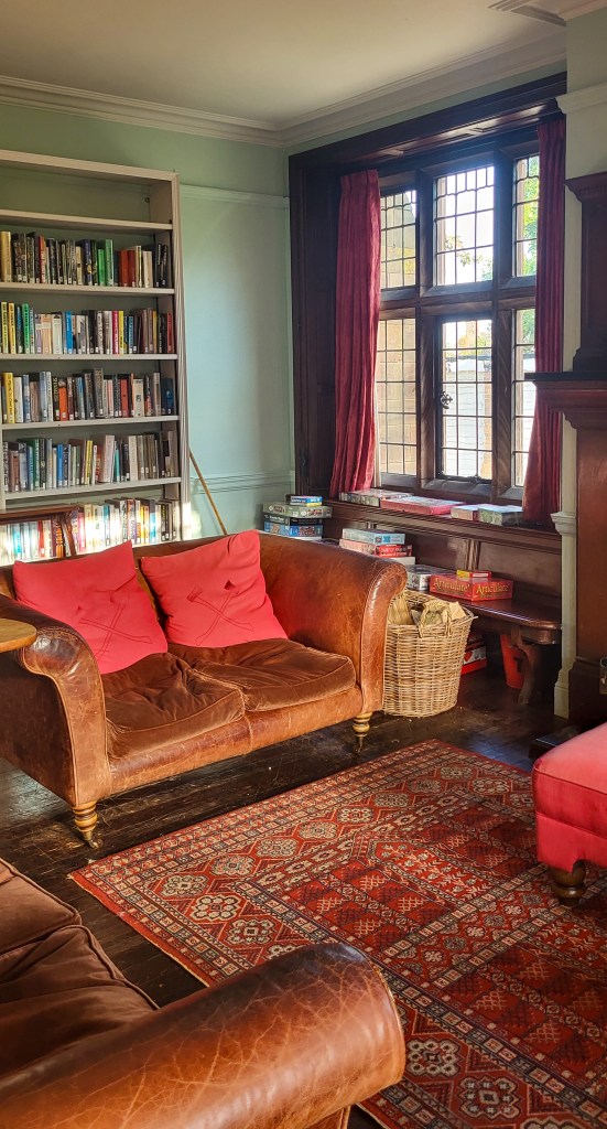 A worn leather sofa with red cushions sits to the right of a Victorian style window. Behind it is a white bookshelves with books. There is a red Persian style rug in front of the sofa.