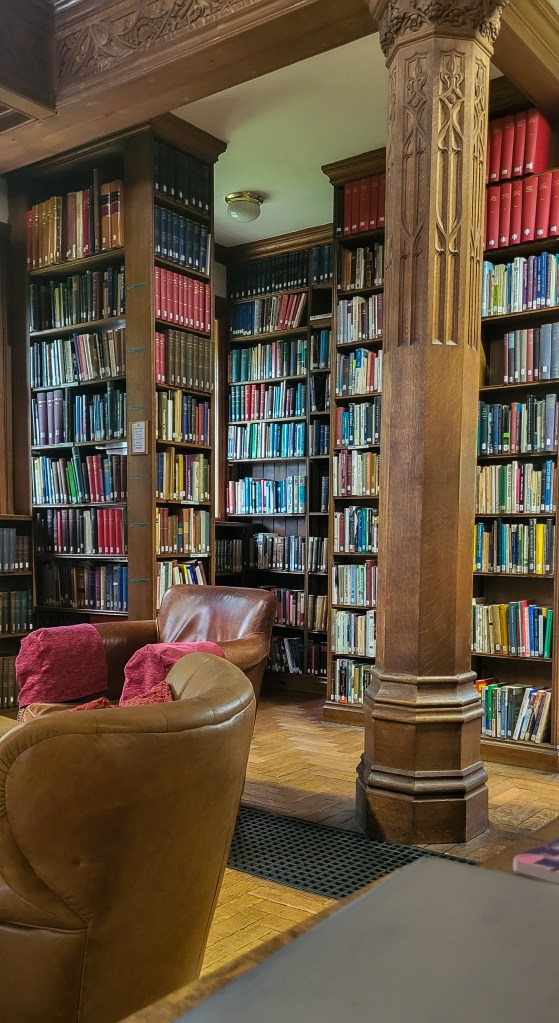 Lines of wooden bookshelves covered in books. To the right of the image is an ornate wooden pillar. To the left of the image are leather comfy armchairs. 