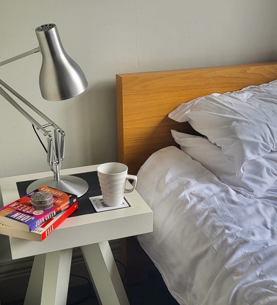 A close up of one of the beds in Gladstone's Library. There is a white bedside table on the left of the image, with a silver reading lamp, a white mug, and two colourful books on it. The right of the images has a wooden bed frame with white pillows and a duvet on it. 