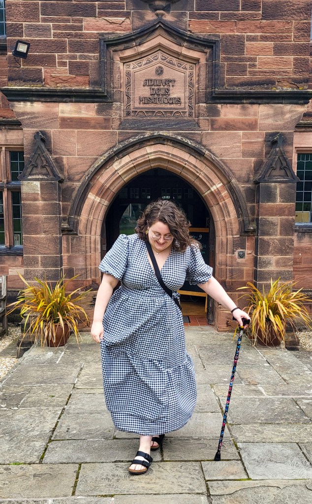 An image of Rosie (a white woman with hard brown wavy hair and glasses) in front of the entrance to Gladstone's. She is wearing a white and black checked midi dress, is looking at the ground, and is holding a black cane in her left hand. There is an archway behind her. 