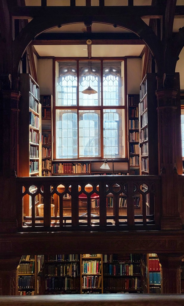 The view of a desk across the mezzanine in Gladstone's library reading room. There is a triple styled window casting sun over the desk that sits between two heavy laden wooden bookshelves.