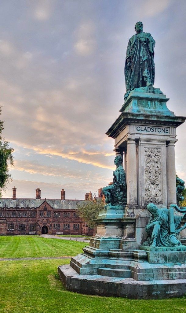In the foreground is a blue and white statue of William Gladstone. He is atop a plinth with his name below him, and there are other blue statues of people at the bottom of the plinth. Behind, you can see the brown brick building of Gladstone's with a white, blue, and orange sky above it, and a square of green grass to the front.