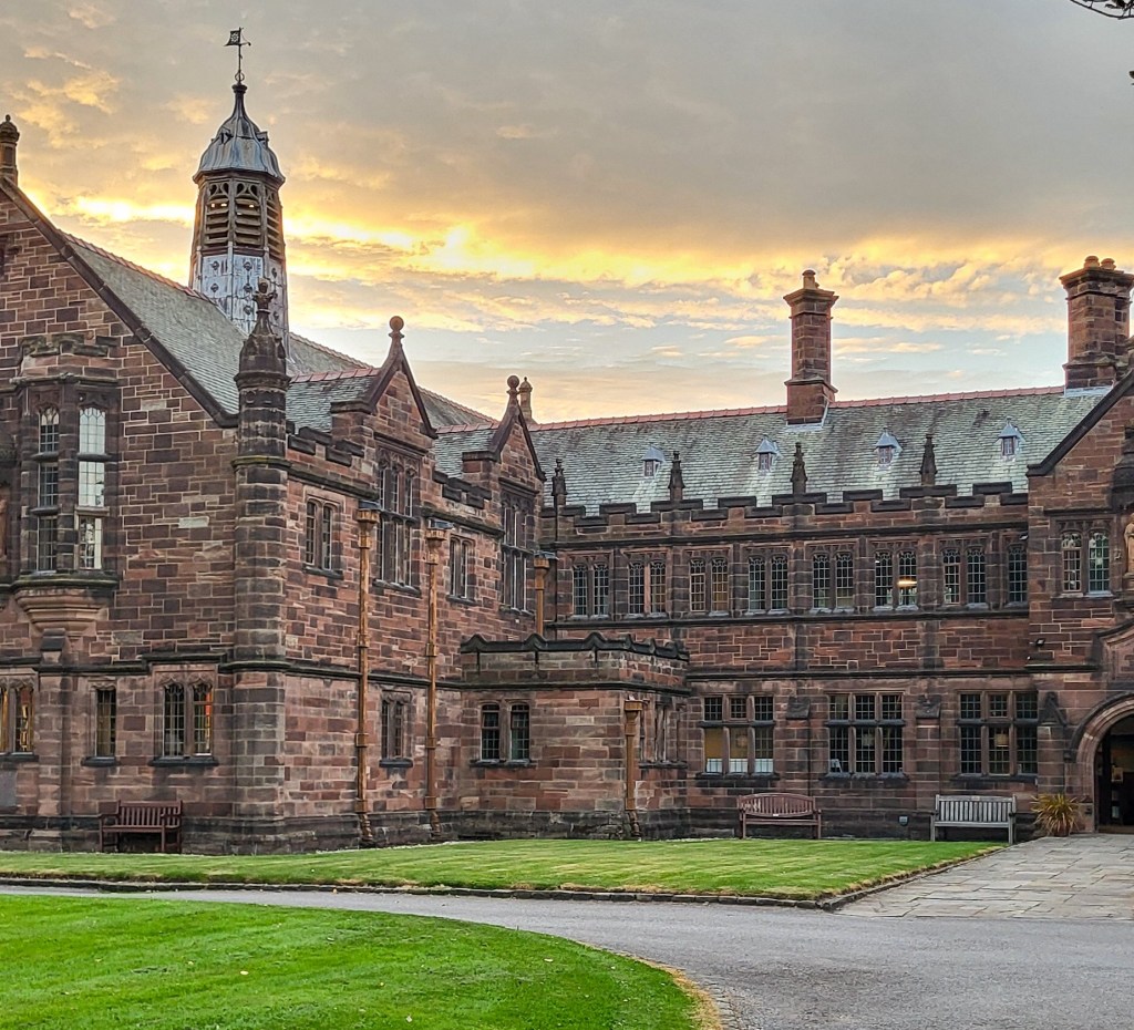A large brown brick building in an L shape, over three floors. There is a square of green grass in front of it. Above, the sky is reflecting a sunset and is white, orange, and blue. 