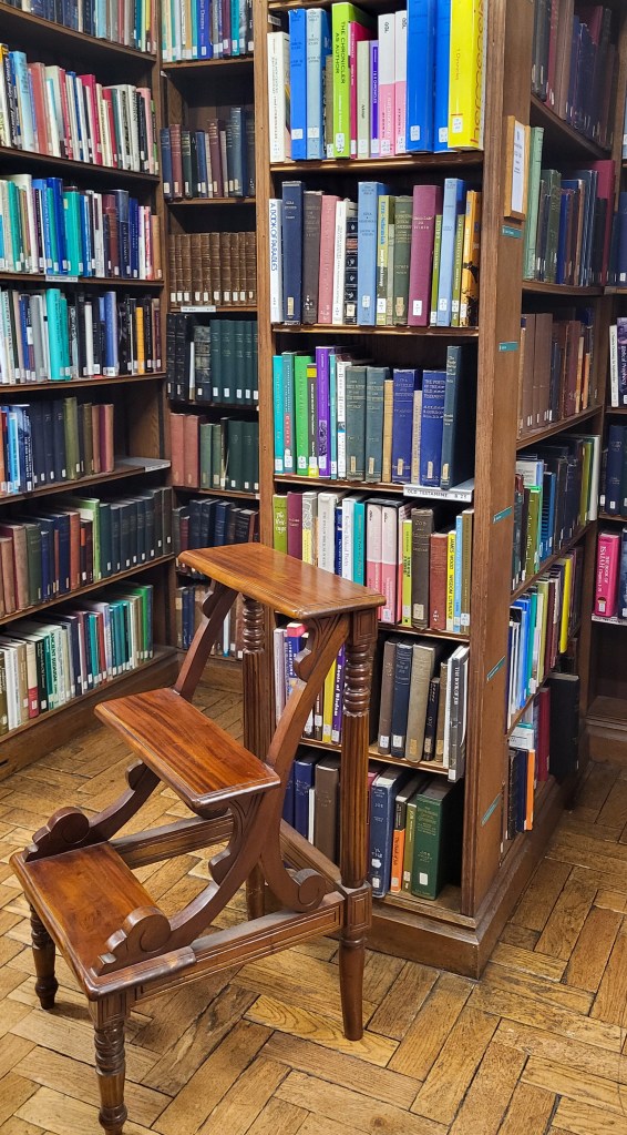Lines of wooden bookshelves covered in books. There is a wooden step stool up against on the of the bookshelves. 