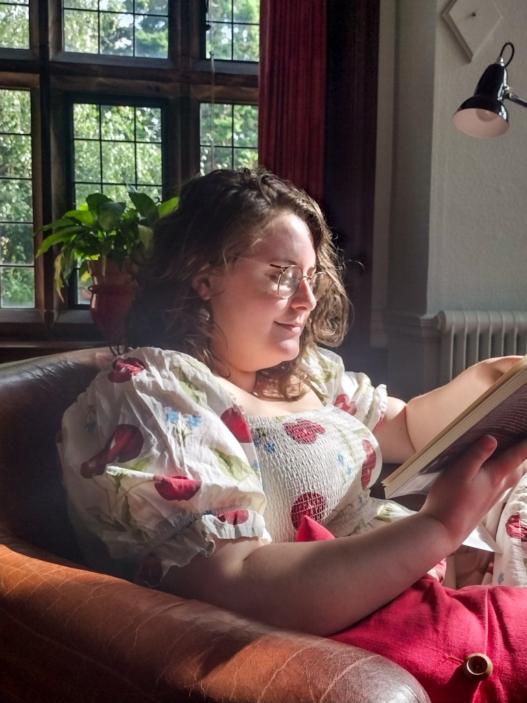 An image of Rosie, the author, reading a book in the Gladstone Room. She is a white woman with wavy brown hair and glasses, and is wearing a cream puff sleeve dress with cherries on it. She is sat in a brown arm chair with red curtains and a window behind her.  