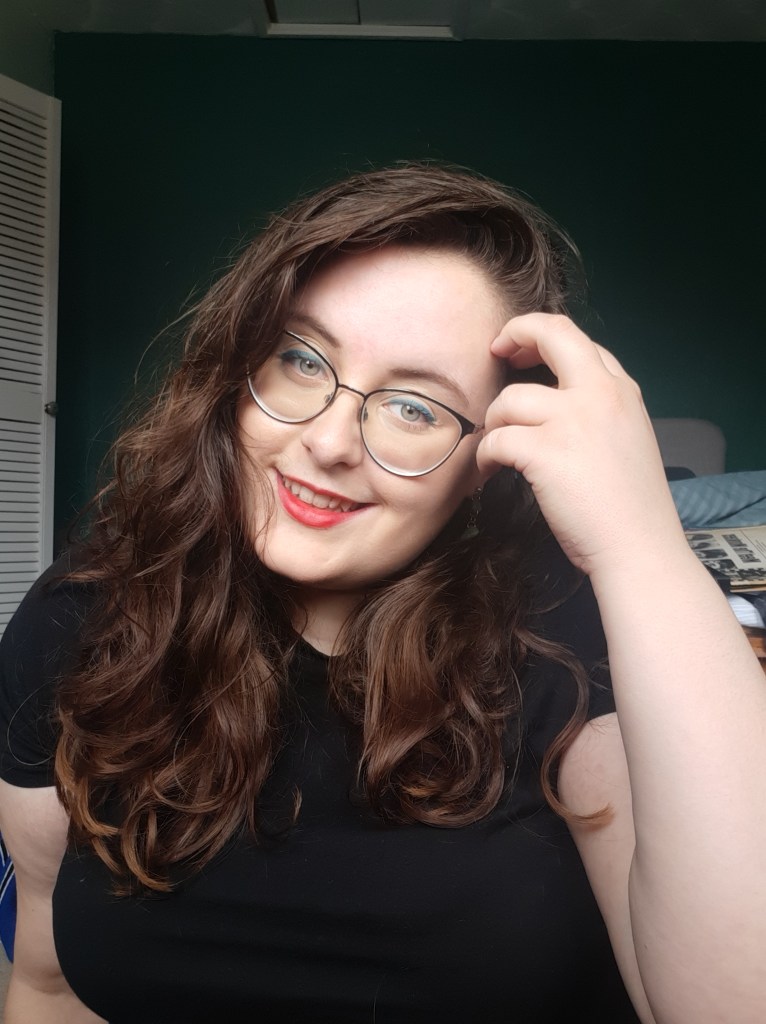 An image of Rosie (a white woman with brown long wavy hair and glasses) smiling into the camera. 