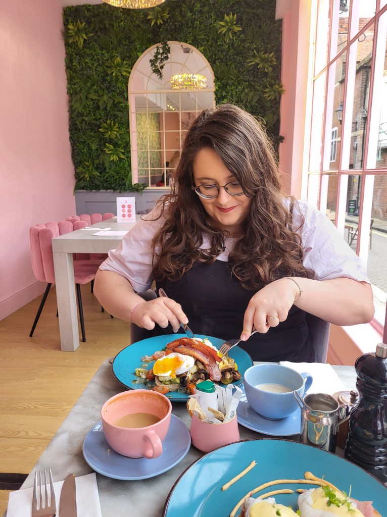 An image of Rosie in a pink cafe, eating a brunch. She is a white woman with glasses, long brown wavy hair, and she is smiling at the plate of food.
