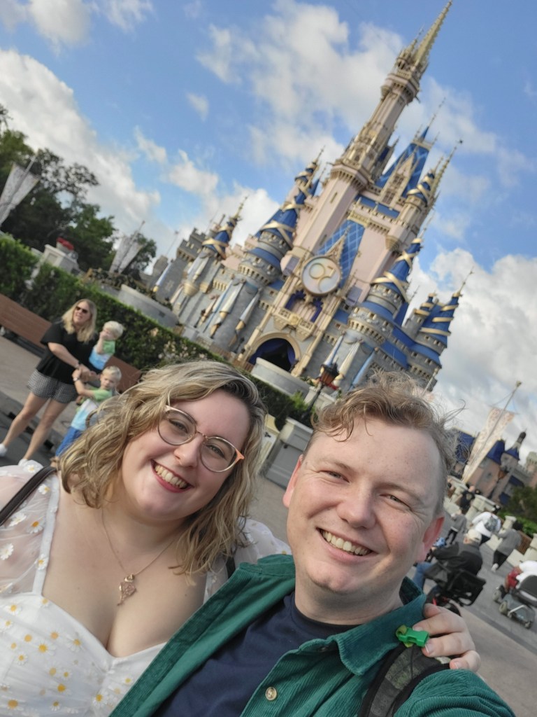 An image of Rosie (a white woman with short blonde hair) and Rory (a white man with quiffed blonde hair) standing in front of Magic Kingdom castle. This is a selfie.