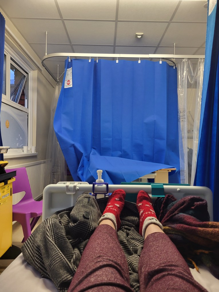 An image of a hospital ward. Rosie's legs are on a bed in red socks. Opposite her is a blue hospital curtain covering another bed.