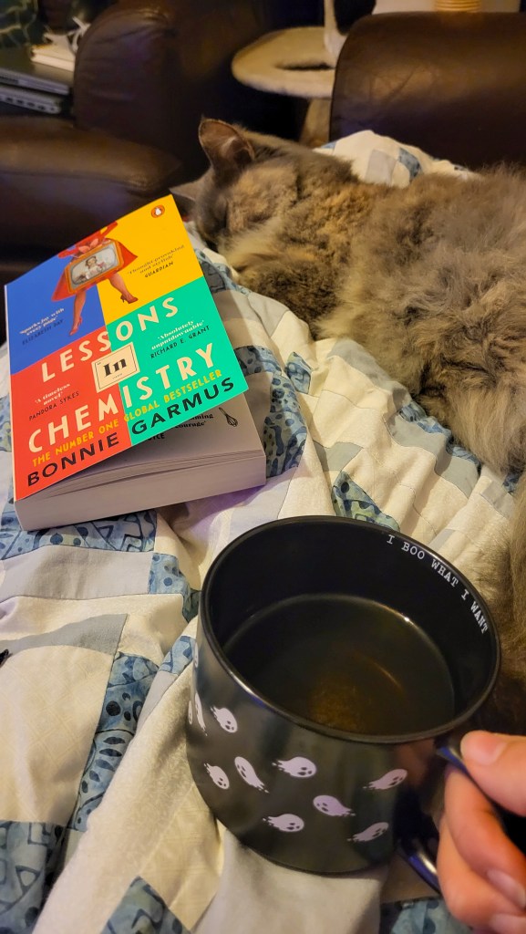 An image of Rosie's evening. Her legs are wrapped in a blue and white blanket, and there is a grey curled up on the blanket. Next to the cat is the book 'Lessons in Chemistry' and below that is a black mug full of tea. 