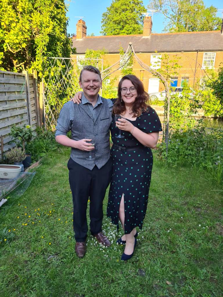 An image of Rory (a white man with blonde quiffed hair) and Rosie (a white woman with brown wavy hair and glasses). They are stood in a garden with a river behind them, both holding wine glasses, and smiling. 
