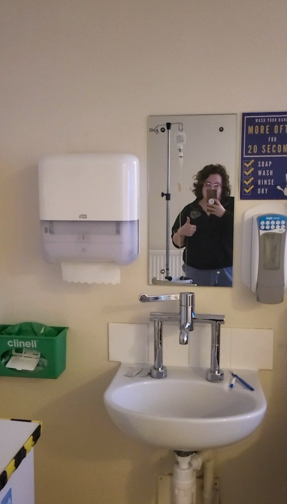 Rosie taking a selfie in a hospital room mirror. She is doing a thumbs up an has an IV next to her.