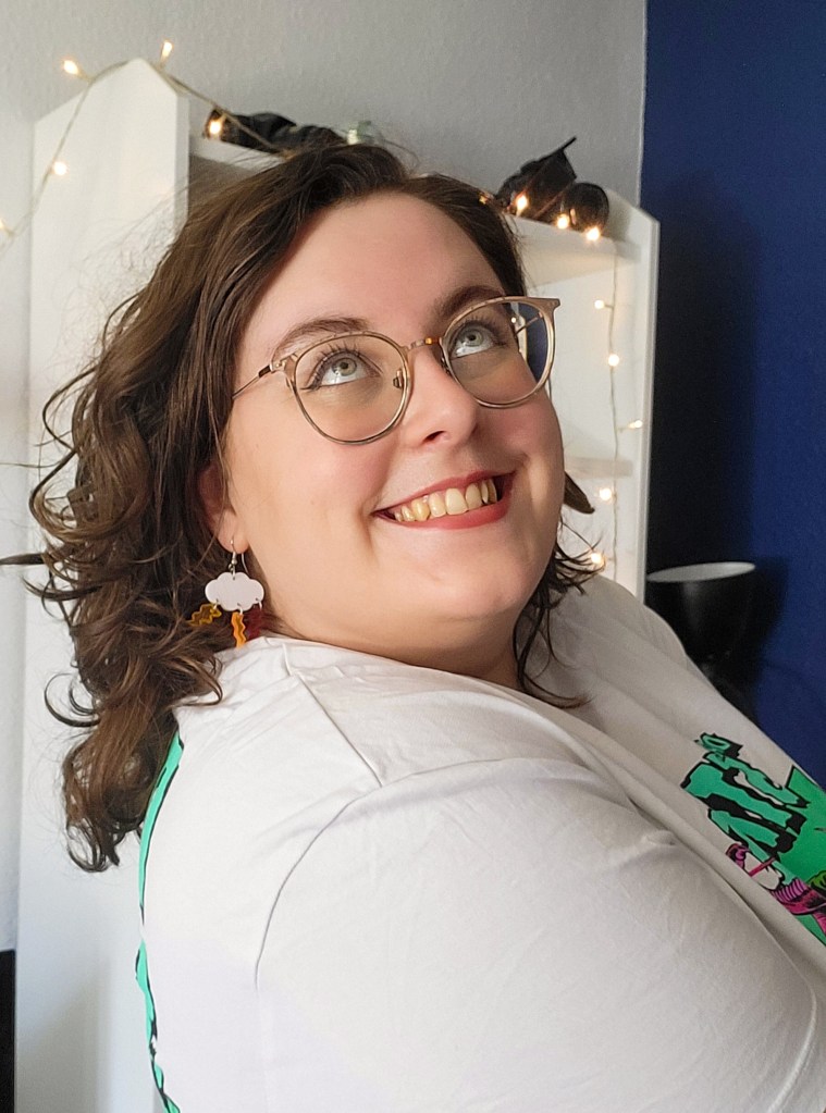 An image of an older, adult Rosie. She has shoulder length wavy hair and glasses, and is looking up to the sky. She is smiling, and wearing 60s inspired make up and a white t shirt. 