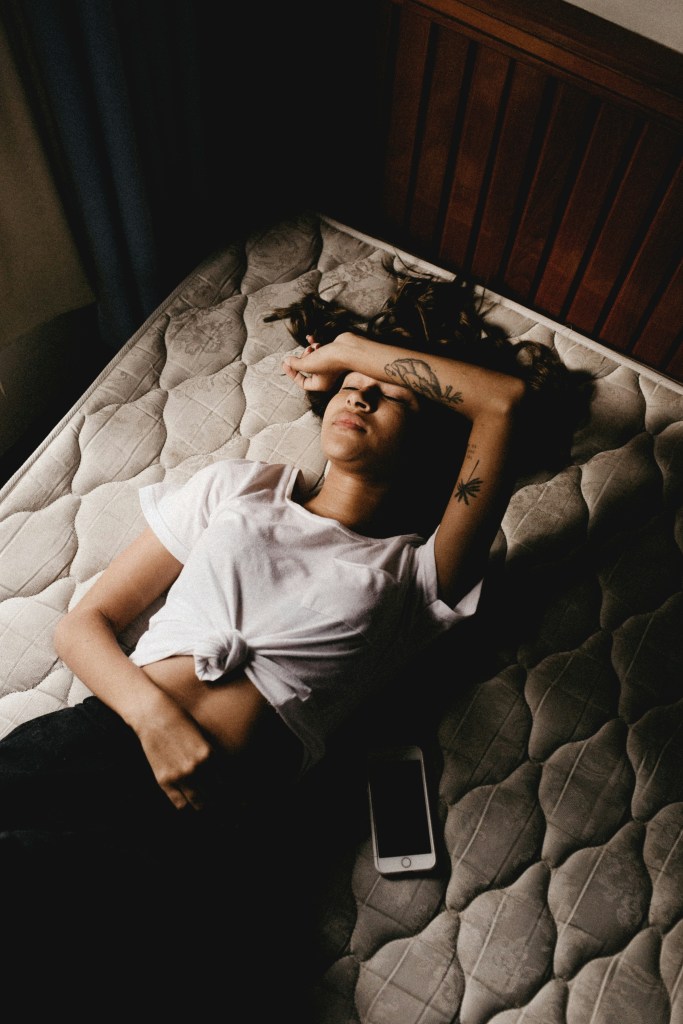 An image of a woman with dark hair laying on a mattress. Her left arm is over her forehead, and her right arm is over her stomach. She has tattoos on her left arm, is wearing a white tee, and black trousers.