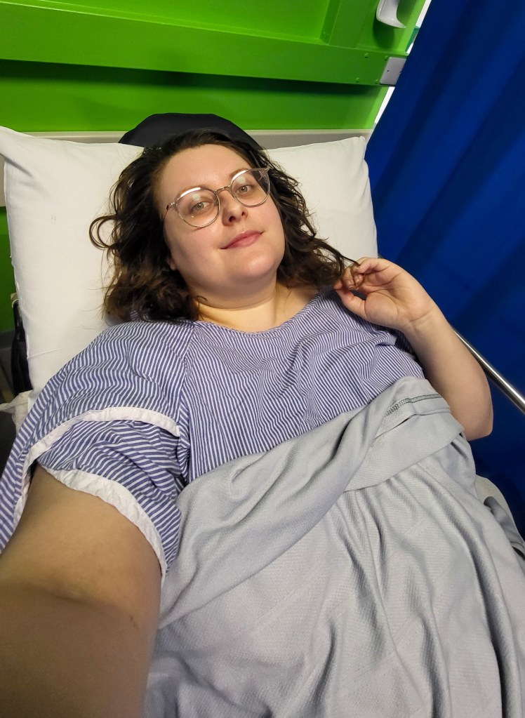 An image of Rosie in a hospital bed. She has brown hair, glasses, white skin, and looks tired. She is wearing a blue and white hospital gown and blanket.