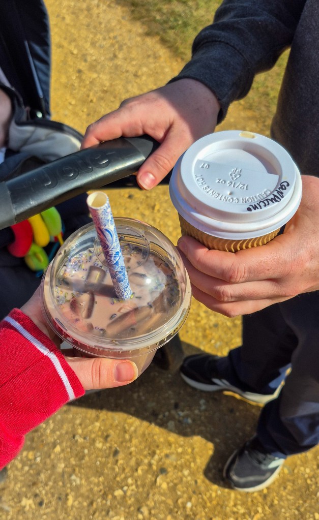 AN image of two hands holding drinks together. On the left is iced coffee in a clear containers with a blue straw. On the right is a brown coffee cup with a white lid, with 'cappuccino' written on the lid.