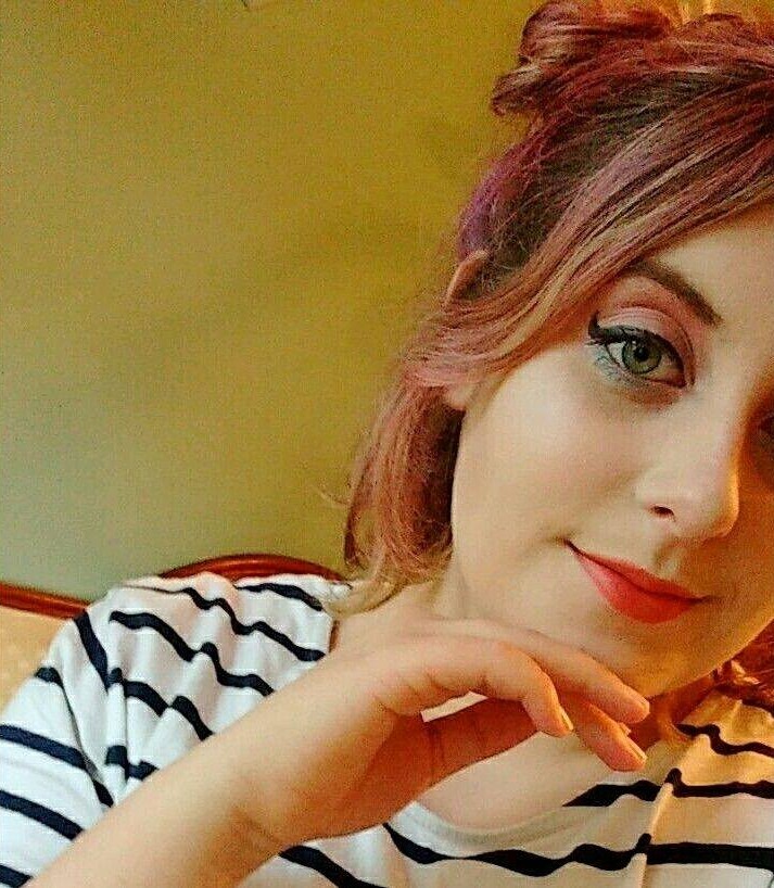 A selfie of Rosie when she was twenty. She has short, pink hair in space buns and thick black eyeliner. She is wearing a striped tee and has her hand on her face. You can only see one eye.