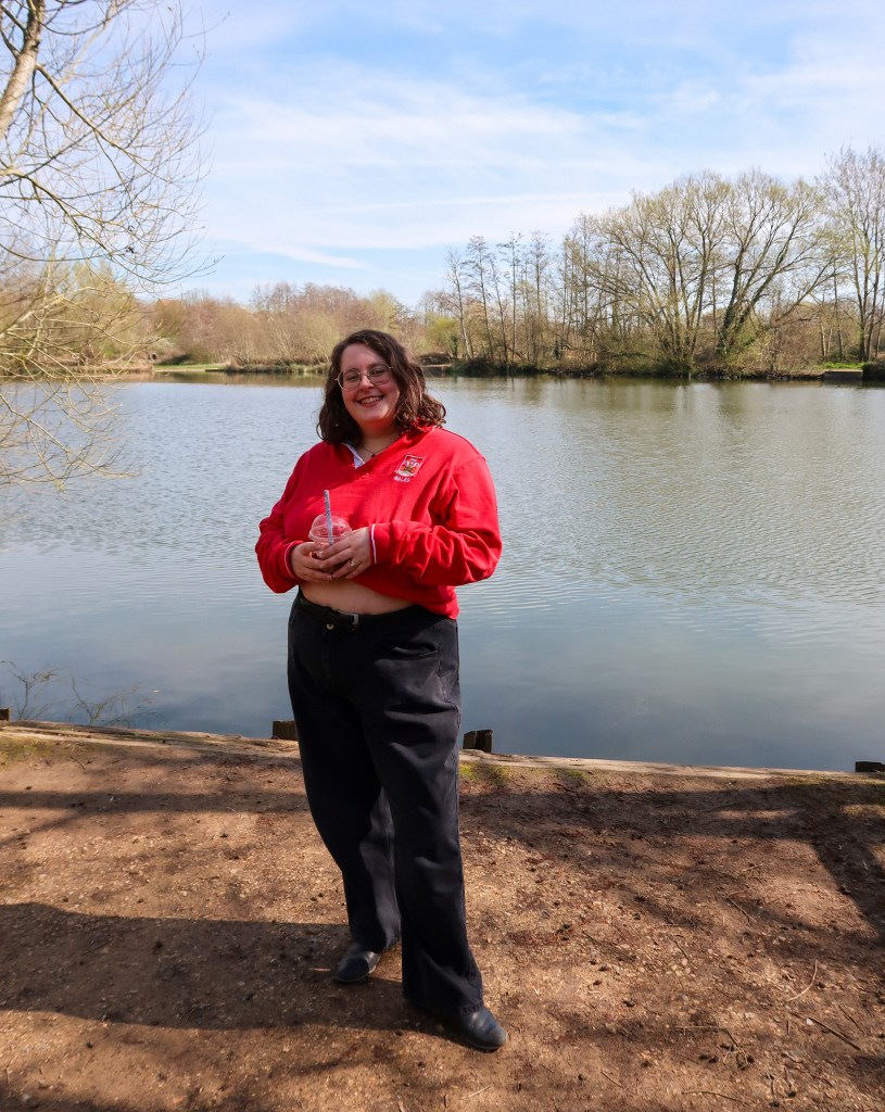 An image of Rosie stood in front of a lake and trees. Rosie is a white woman with wavy brown hair and glasses. She is wearing a red rugby shirt and black jeans, and is holding an iced coffee in her hands. She is smiling at the camera with a foot popped.