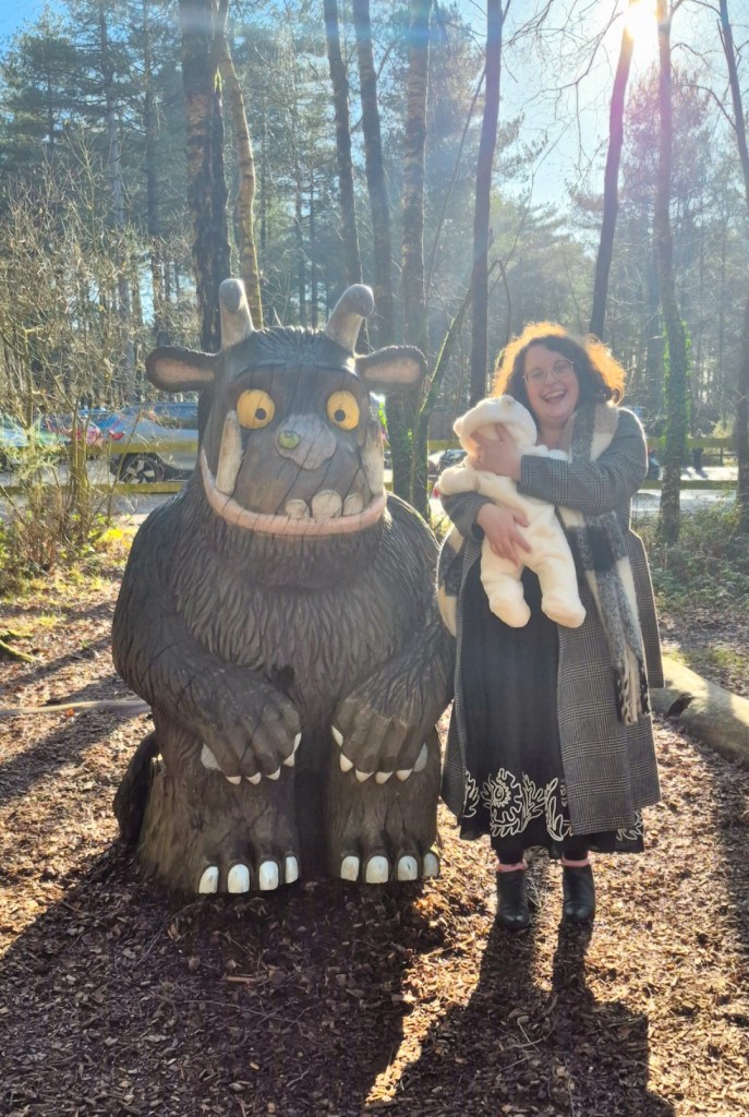An image of Rosie and a baby in an all-in-one coat next to a statue of the Gruffalo. Rosie is wearing a black dress and grey coat, has brown hair and glasses, and is smiling at the camera. The baby is in her arms, back to the camera.